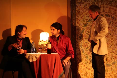 A scene from Pezzi Scomposti: two men sitting at a small table with a lamp, a third one standing